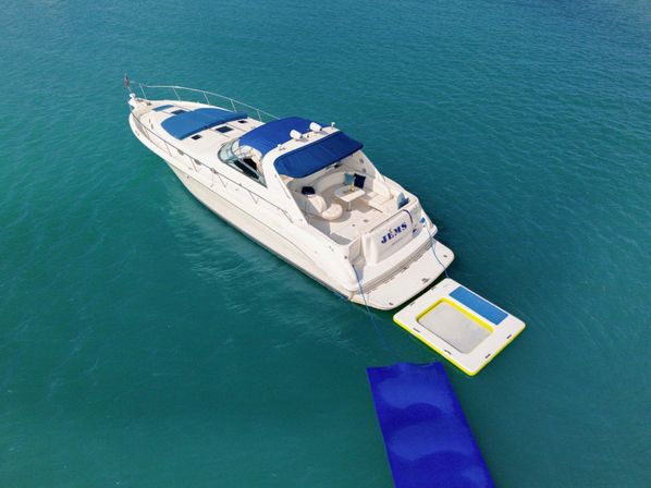 Aerial view of a white luxury motor yacht floating in clear turquoise water with blue sun awnings, an open aft lounge, and an attached inflatable swim platform and blue floating ramp.