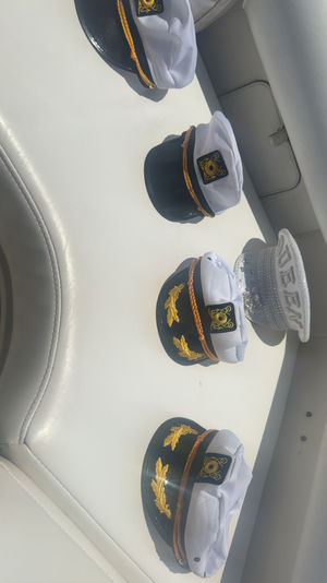 Three black-and-white captain hats with gold trim and nautical insignia plus a sparkly silver party top hat arranged on a white leather boat bench in bright sunlight — playful nautical scene.
