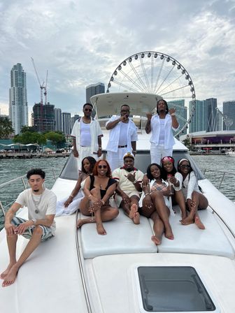 Group of friends lounging on the bow of a white yacht with the Miami downtown skyline and a large waterfront Ferris wheel under a cloudy sky
