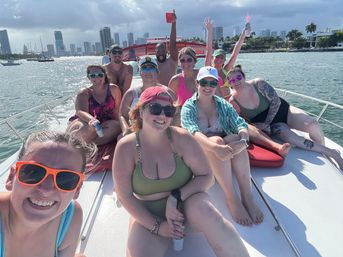 Smiling group of people in swimsuits lounging on the bow of a boat in sunny Miami bay, holding drinks with the city skyline and palm trees in the background.