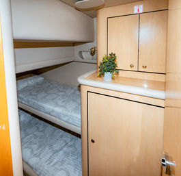 Small yacht cabin with stacked bunk beds, pale wood cabinetry, brass reading light and a potted plant on the counter