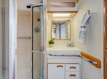 Compact RV/boat-style bathroom with frosted glass corner shower, white vanity and sink, framed mirror, chrome grab bar, striped hand towel and small potted plant.