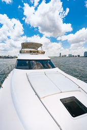 Front view of a white luxury yacht bow with cushioned sun pads and glass hatch, cruising in a calm harbor with a distant coastal skyline of high-rise buildings under a bright blue sky dotted with fluffy clouds.