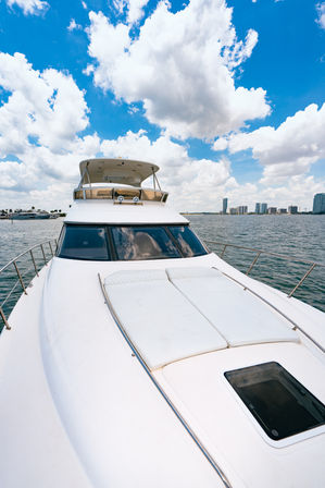 Front view of a white luxury yacht bow with cushioned sun pads and glass hatch, cruising in a calm harbor with a distant coastal skyline of high-rise buildings under a bright blue sky dotted with fluffy clouds.