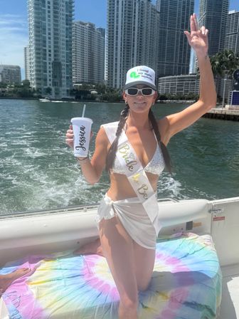 Bachelorette boat party scene: bride-to-be in a white bikini and sash holding a drink and waving, with a sunny coastal city waterfront and tall high-rise buildings in the background.