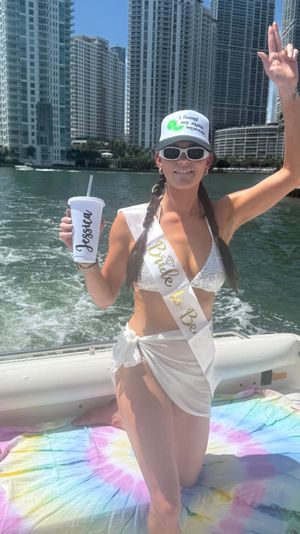 Bachelorette celebrating on a boat — woman in white bikini and 'Bride to Be' sash wearing sunglasses and a cap, holding a drink with a city waterfront high-rise skyline and tie-dye towel in the sun.