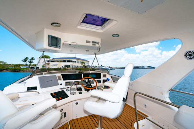 Sunlit yacht helm with white leather captain’s chair, wooden steering wheel and navigation panels, overlooking turquoise marina waters, waterfront buildings and a distant bridge under a bright blue sky.
