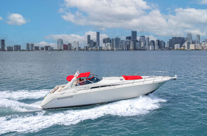 Sleek white luxury motor yacht with red sunshades slicing through turquoise waters with Miami skyline and fluffy clouds in the background