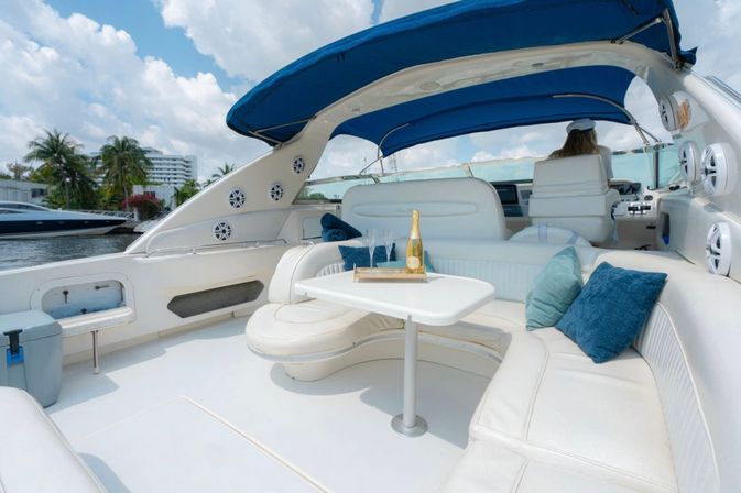 Sunlit luxury yacht lounge at a tropical marina with white curved leather seating, blue pillows, a table holding champagne and flutes, and a blue bimini top