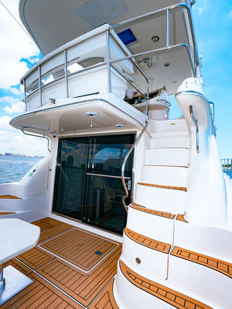 Luxury white yacht aft with teak deck and curved staircase to the upper deck, stainless railings, glass sliding door and coastal skyline on a sunny day.