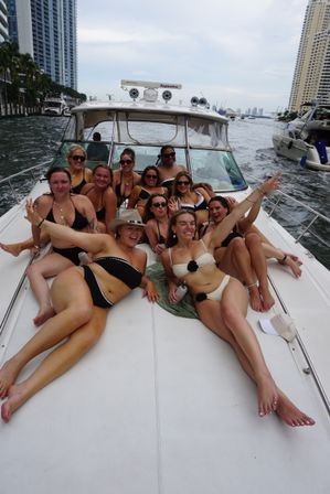 Bikini-clad friends lounging on the bow of a luxury yacht cruising an urban waterway, high-rise skyline and other boats in the background for a lively daytime boat party vibe.