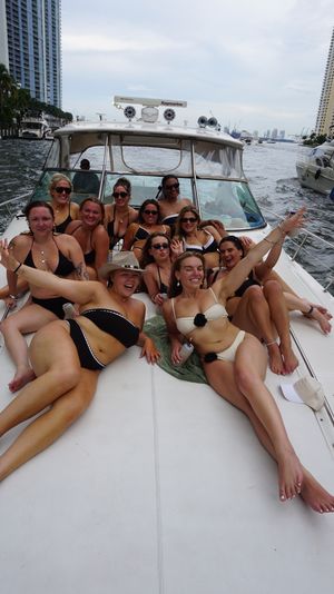 Group of women in bikinis lounging and celebrating on the bow of a motor yacht cruising a downtown waterfront canal lined with high-rise buildings and other boats