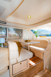 Sunlit yacht interior with cream leather wraparound seating and a small white dining table, wood flooring, window blinds framing palm trees and marina view, potted plant and model sailboat on the ledge.