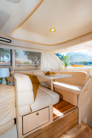 Sunlit yacht interior with cream leather wraparound seating and a small white dining table, wood flooring, window blinds framing palm trees and marina view, potted plant and model sailboat on the ledge.