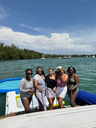 Five women in swimsuits pose on a boat swim platform with paddles and an inflatable mat in a turquoise coastal inlet, anchored boats and mangrove shoreline under a bright blue sky.