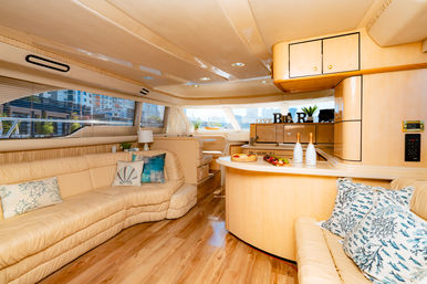 Sunlit luxury yacht salon with curved beige leather sofas, wood floors, compact bar counter with champagne and fruit, decorative pillows and large windows overlooking a marina and waterfront city skyline.