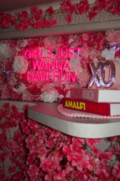 Pink neon sign reading "GIRLS JUST WANNA HAVE FUN" on a lush pink floral wall with stacked decor books and a glittery XO accent — playful, Instagrammable party photo backdrop.