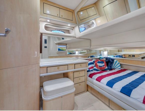 Cozy luxury yacht cabin interior with light wood finishes, nautical red-white-blue striped bedding, built-in drawers and cabinets, vanity with cushioned stool, porthole window and mirrored walls.