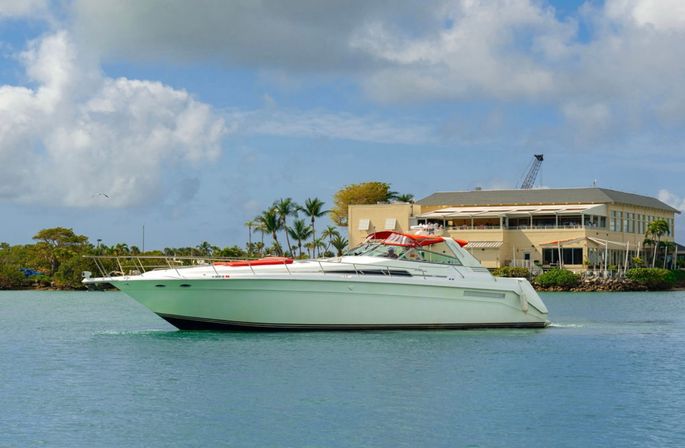 Gleaming white motor yacht cruising on turquoise water past a tropical marina with palm trees and a waterfront building under a blue sky with puffy clouds.