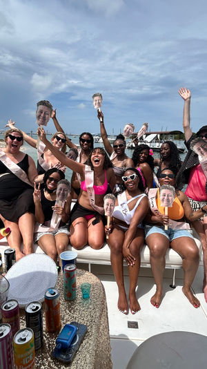 Bachelorette party on a boat: a lively group in swimsuits and sashes cheering on deck near a coastal beach and anchored boats, waving oversized face cutouts under a bright summer sky with drinks on the table.