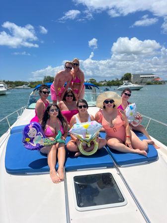 Group of friends in colorful swimsuits lounging on a yacht bow in a sunny coastal marina, holding inflatable diamond-ring and mermaid balloons.