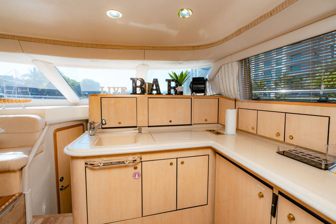Bright luxury yacht galley with light wood cabinets and countertops, small sink, decorative "BAR" letters, paper towel holder, window blinds and marina view.