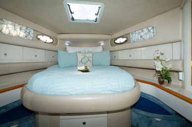 Cozy yacht v-berth cabin with round bed dressed in light-blue linens, decorative sea-turtle pillow, overhead hatch and oval portholes, built-in storage cabinets and orchid accent.