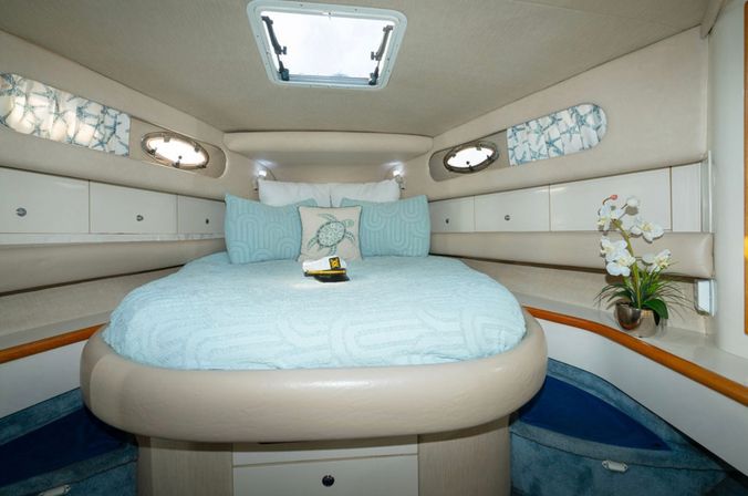 Cozy yacht v-berth cabin with round bed dressed in light-blue linens, decorative sea-turtle pillow, overhead hatch and oval portholes, built-in storage cabinets and orchid accent.