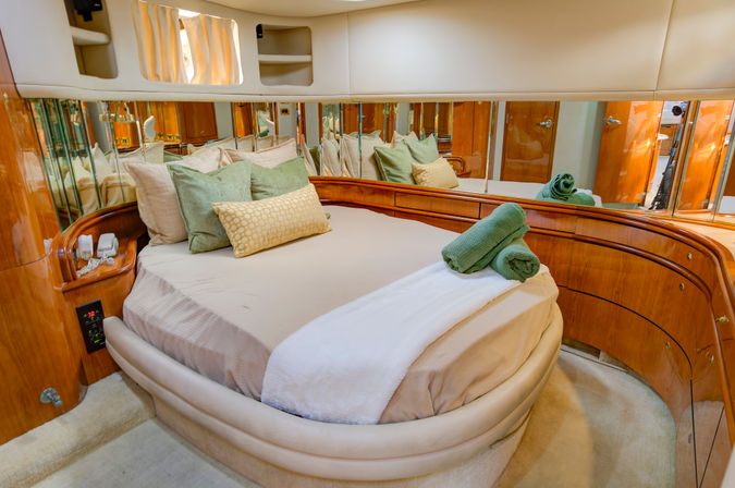 Cozy luxury yacht master stateroom with rounded bed, mirrored walls, glossy wood cabinetry, green and beige pillows and rolled towels.