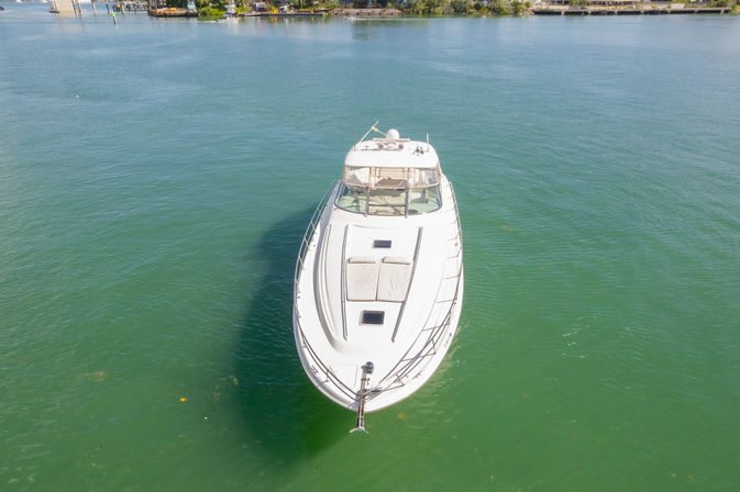 Aerial photo of a sleek white luxury yacht floating in clear turquoise coastal harbor water near a marina.