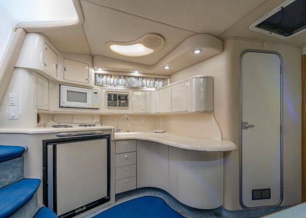Cozy cream-colored yacht galley with curved cabinets, microwave, sink, mini-fridge, recessed LED lighting and blue-carpeted steps in a compact boat cabin