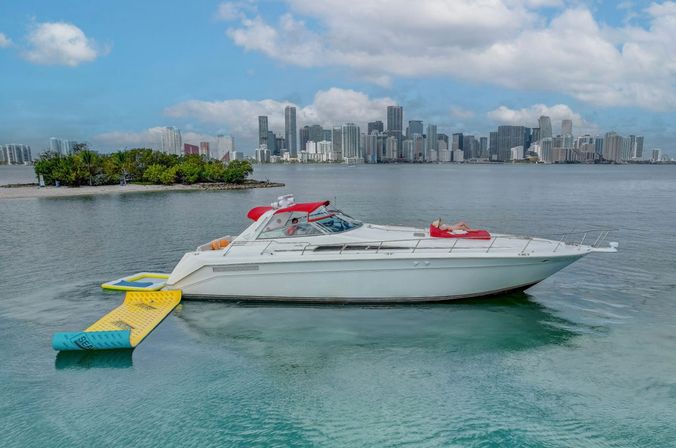 White luxury yacht anchored in turquoise bay with yellow-blue inflatable swim platform and a sunbather on the bow, downtown Miami skyline and a small island in the background.