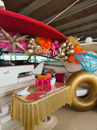 Bachelorette party setup aboard a yacht with gold, pink and orange balloon garland, Bride to be banner, giant gold ring float, sequin gold tablecloth and hot-pink champagne flutes