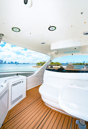 Sunny yacht cockpit with teak-style deck and curved white seating, panoramic view of turquoise bay, small tree-covered islet and distant city skyline under a bright blue sky.
