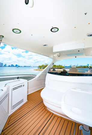 Sunny yacht cockpit with teak-style deck and curved white seating, panoramic view of turquoise bay, small tree-covered islet and distant city skyline under a bright blue sky.