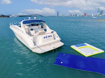 White luxury yacht moored in turquoise water with a blue swim mat and neon-trim inflatable platform tied nearby, urban skyline and a bridge on the horizon under a sunny sky.