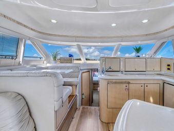Bright luxury yacht interior salon and galley with white leather seating, light wood cabinetry, curved countertops and panoramic windows framing blue ocean and sky views.