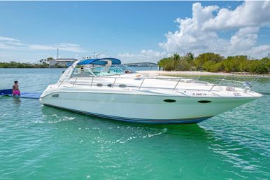 White luxury motor yacht anchored in turquoise water off a sunny Florida coast, mangrove-lined shore and small bridge in the background, with a person on a blue paddle mat beside the boat.