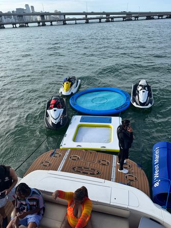 View from a yacht swim platform showing three jet skis and a round blue floating trampoline tied alongside, guests lounging on the deck, and a distant city skyline with a highway bridge over the water.
