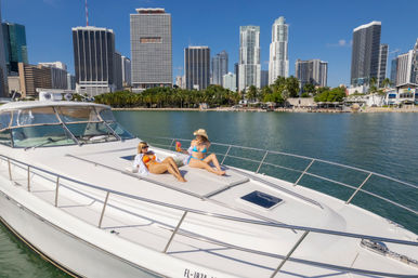 Two women sunbathing on the bow of a white yacht in Miami Bay, sipping colorful drinks with downtown Miami skyline, palm trees, and clear blue sky in the background.