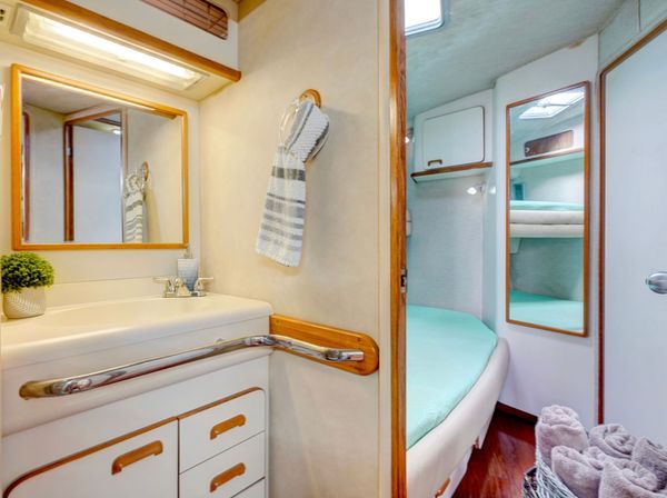 Cozy boat interior: compact yacht bathroom with sink, mirror, striped hand towel and chrome grab rail opening to a snug cabin with a mint-green berth, mirrored storage and a basket of rolled towels.