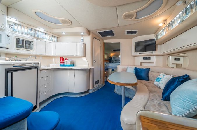 Light-filled yacht salon and galley with vibrant blue carpet, curved cream leather sofa, wood-trim table, white cabinets, TV and sleeping cabin ahead.