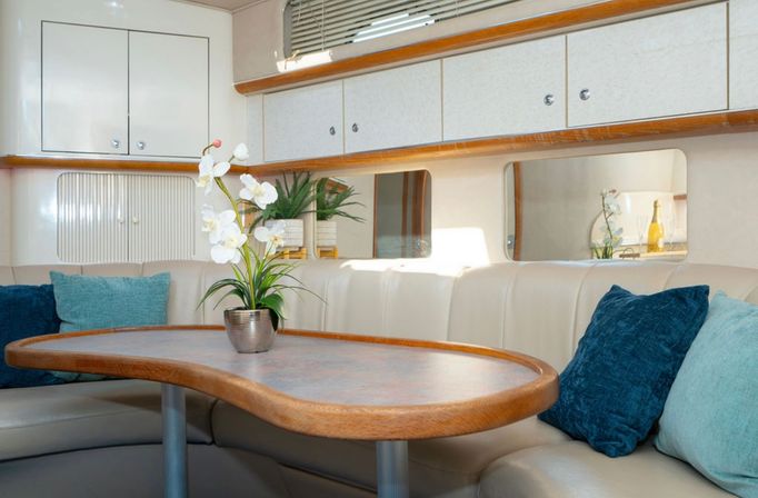 Cozy yacht cabin dining nook with curved wood-trimmed table, potted white orchid centerpiece, cream leather banquette and teal throw pillows under overhead cabinets