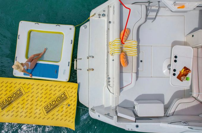 Aerial view of a white motorboat tied to a yellow inflatable dock and square floating lounge in turquoise water, a sunbather in a white swimsuit and straw hat reclines on the float, striped towel and orange cushions on the boat with drinks on the table — summer boating scene.