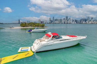 White motor yacht with red canopy anchored by a yellow floating ramp near a small sandbar in turquoise waters, Miami skyline and puffy clouds in the background.