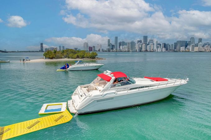 White motor yacht with red canopy anchored by a yellow floating ramp near a small sandbar in turquoise waters, Miami skyline and puffy clouds in the background.