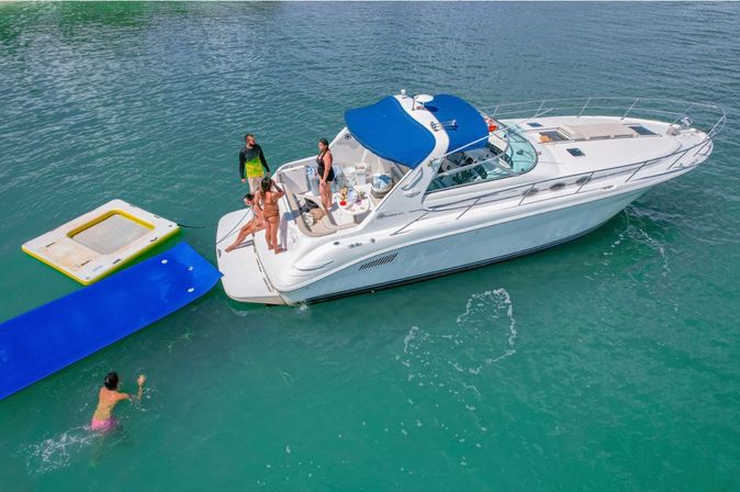 White motor yacht with blue canopy anchored in turquoise coastal water, people on the swim platform with an inflatable trampoline and blue water mat while someone swims nearby