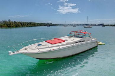 White luxury motor yacht with red sun pad anchored in clear turquoise tropical bay, sailboats and tree-lined shoreline under a sunny blue sky.