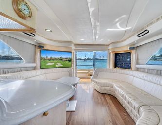 Sunlit luxury yacht salon with curved cream leather seating, wood floors, TV showing a golf course, nautical clock and control panel, sliding glass doors opening to a blue marina and harbor cranes in the distance.