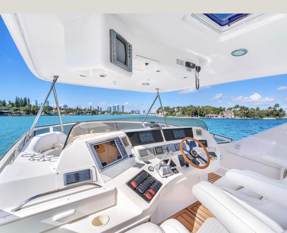 Luxury yacht helm on a sunny day — wooden steering wheel, multi-screen navigation console and white leather seats overlooking turquoise water and a palm-lined coastal skyline.